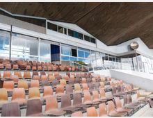 Gradinate di uno stadio o arena con file di sedili in plastica marrone disposti su più livelli. Sullo sfondo si vede una struttura coperta con grandi vetrate, ringhiere metalliche e un soffitto inclinato in cemento.