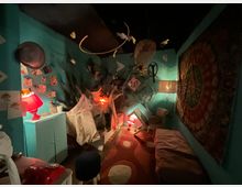 Interno di una piccola stanza scenografica con pareti turchesi, illuminata da lampade rosse e con mobili e oggetti inclinati come in un’installazione. Sul fondo c’è un effetto “esplosione” o squarcio scuro, con decorazioni sospese al soffitto e un grande pannello decorativo sulla parete destra.