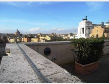 Terrazza panoramica con muretto in pietra e fioriere, affacciata su un ampio skyline urbano. Sullo sfondo si distinguono cupole e un grande edificio monumentale bianco con colonnato e statua in sommità.