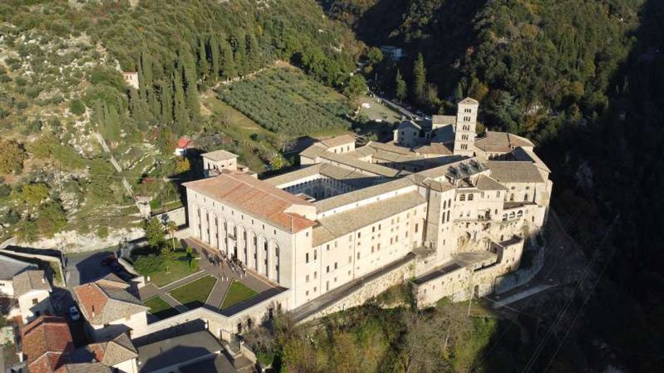 Veduta aerea di un grande complesso monastico in pietra con tetti in coppi rossi, chiostro interno e campanile, affacciato su una scarpata. Intorno si vedono una valle boscosa, campi coltivati e piccole strade di accesso.