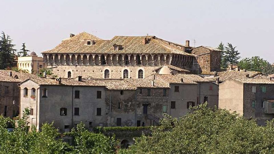 Veduta panoramica di un centro storico con edifici in pietra e tetti in coppi, dominato da un grande palazzo antico con loggiato ad archi e copertura a falde. In primo piano si vedono alberi e vegetazione che incorniciano le case addossate tra loro.