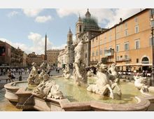 Fontana monumentale con statue in marmo in una grande piazza urbana, con acqua in vasca e sculture di figure mitologiche. Sullo sfondo si vedono edifici storici e una chiesa con cupola e impalcature, oltre a un obelisco al centro della piazza.