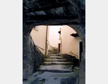 Vicolo stretto in un borgo storico, con gradini in pietra e pavimentazione irregolare che salgono tra muri in intonaco chiaro. La scena è incorniciata da un arco basso con travi di legno scure, con una ringhiera metallica e piccole porte e finestre sulle facciate.