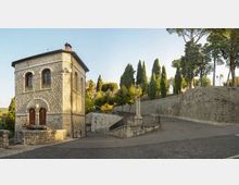 Edificio in pietra a due piani con archi e finestre ad arco, affacciato su una strada in salita. Accanto si vedono mura in pietra, una scalinata con ringhiera e alberi alti (cipressi e pini) lungo il pendio.