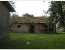 Piccola casa rurale in muratura con tetto a due falde coperto di tegole e muschio, con ingresso ad arco e finestre con inferriate. Davanti si estende un prato verde, con alberi ai lati e un altro edificio parzialmente visibile sulla sinistra.