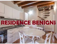Cucina-soggiorno open space con mobili bianchi e piano cottura lineare lungo la parete, tavolo rotondo con sedie al centro e soffitto con travi in legno a vista. Sul lato destro si vede una scala a chiocciola in metallo che conduce al piano superiore.