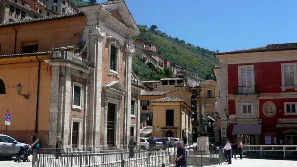 Piazza di un centro storico con una piccola chiesa in pietra con facciata rettangolare, portale centrale e croce sul tetto, circondata da ringhiera metallica. Sullo sfondo si vedono edifici colorati e case sul pendio di una collina verde sotto un cielo sereno.