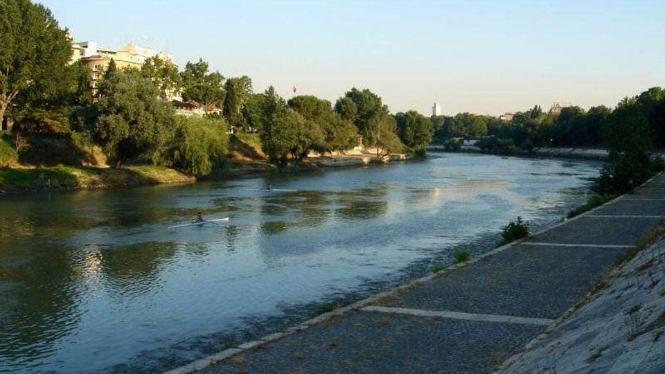 Vista di un fiume che scorre tra sponde alberate, con un’ampia banchina in pietra e una passeggiata lastricata lungo la riva destra. Sullo sfondo si intravedono edifici e vegetazione sotto un cielo sereno.