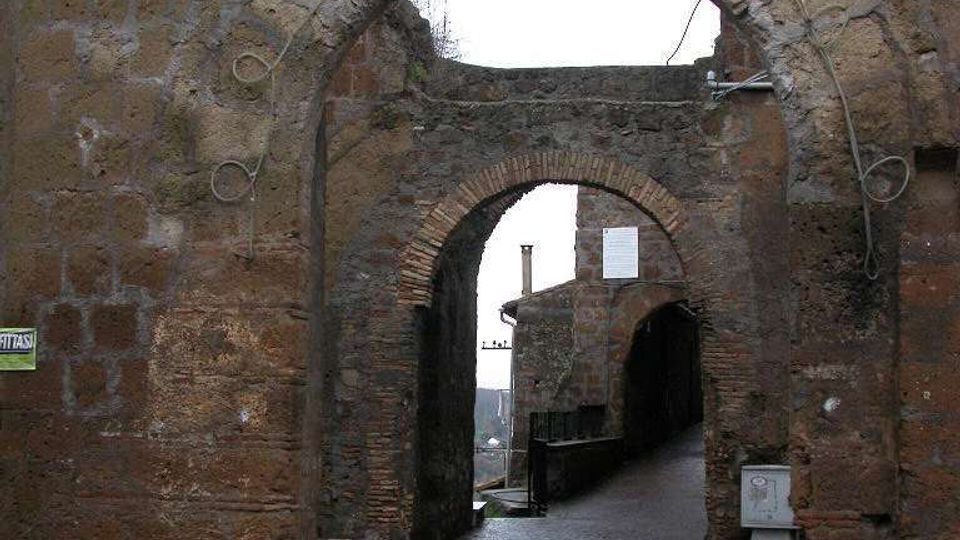 Arco in pietra di una porta medievale con stemma scolpito, che attraversa una spessa muratura in blocchi. Sotto l’arco si vede un secondo passaggio ad arco e una stretta strada lastricata in leggera discesa tra edifici in pietra.