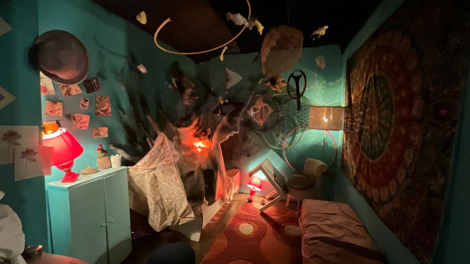 Interno di una piccola stanza scenografica con pareti turchesi, illuminata da lampade rosse e con mobili e oggetti inclinati come in un’installazione. Sul fondo c’è un effetto “esplosione” o squarcio scuro, con decorazioni sospese al soffitto e un grande pannello decorativo sulla parete destra.