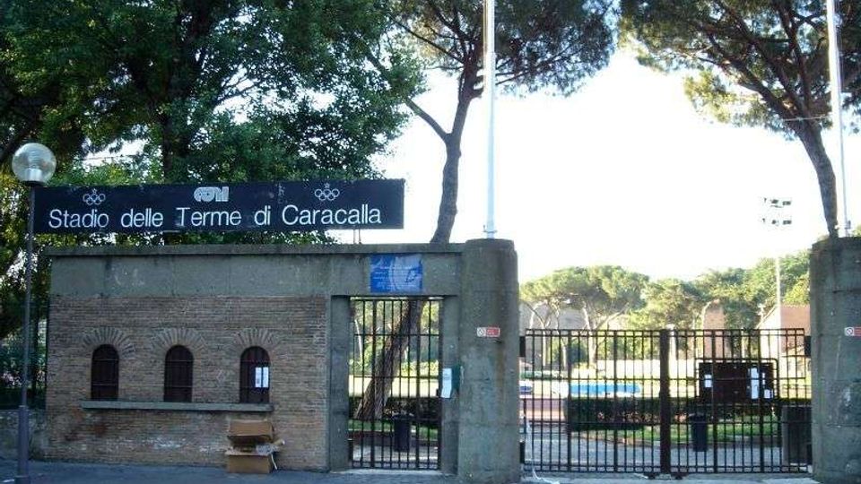 Ingresso dello Stadio delle Terme di Caracalla con cancello in ferro e muri in mattoni e cemento, sormontato da un’insegna con il nome dello stadio. Sullo sfondo si vedono alberi alti e parte dell’area sportiva interna.