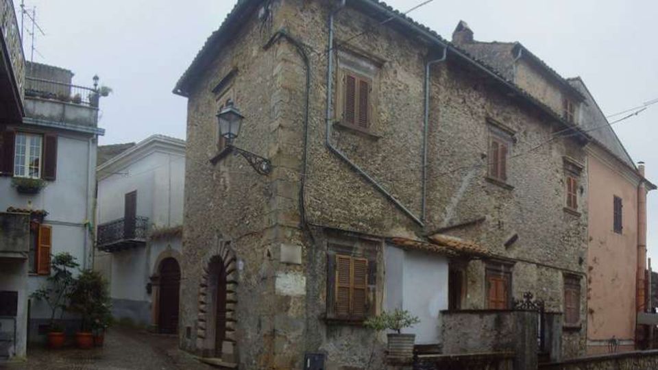 Edificio storico in pietra con intonaco consumato e persiane in legno, su un angolo tra una stradina acciottolata e una piccola piazza. Si notano un arco in pietra al piano terra, una lanterna a muro e una scalinata con muretto e ringhiera sul lato destro.