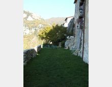 Striscia di prato lungo un edificio in pietra su un pendio, con muretto a secco sul lato sinistro e rocce affioranti vicino alla facciata. Sullo sfondo si vedono colline boscose e montagne.