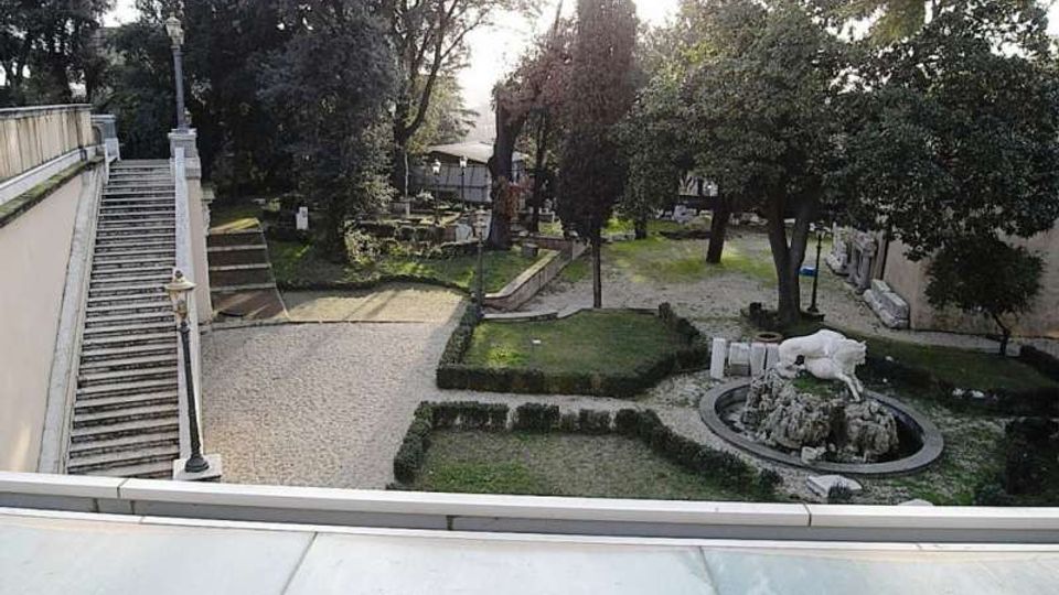 Giardino storico con vialetti in ghiaia e aiuole geometriche delimitate da siepi, circondato da alberi alti. A sinistra una lunga scalinata con balaustra e lampioni, a destra una fontana circolare con una scultura.