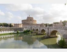 Veduta di Castel Sant’Angelo a Roma, grande fortezza cilindrica in pietra, con il Ponte Sant’Angelo ad archi in primo piano che attraversa il fiume Tevere. Sullo sfondo si vedono mura, edifici storici e alberi lungo le rive sotto un cielo parzialmente nuvoloso.
