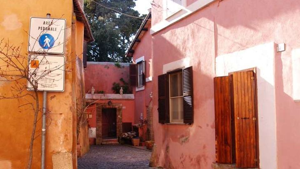 Vicolo pedonale acciottolato tra edifici a due piani intonacati in tonalità rosa e arancione, con persiane e portone in legno. Sulla parete sinistra sono visibili un lampione a muro e un cartello “area pedonale”, mentre il vicolo conduce a un piccolo cortile con ingresso ad arco.