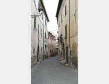 Stradina stretta in un centro storico, pavimentata in sampietrini e fiancheggiata da alti edifici intonacati con persiane verdi e lampioni a parete. Lungo le facciate si vedono portali ad arco in pietra, muri leggermente usurati e cavi sospesi tra le case.