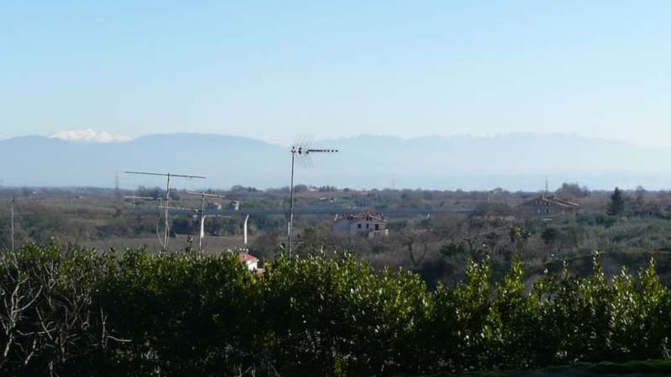 Veduta panoramica di una campagna con vegetazione bassa in primo piano e alcune case sparse e antenne sullo sfondo. All’orizzonte si distinguono colline e una catena montuosa in lontananza sotto un cielo sereno.