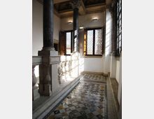 Loggia o corridoio interno di un edificio storico con colonne in pietra e balaustra scolpita, soffitto a cassettoni e grandi finestre con vetri a losanghe e scuri in legno. Pavimento in piastrelle decorate a motivi geometrici, illuminato dalla luce naturale che entra lateralmente.