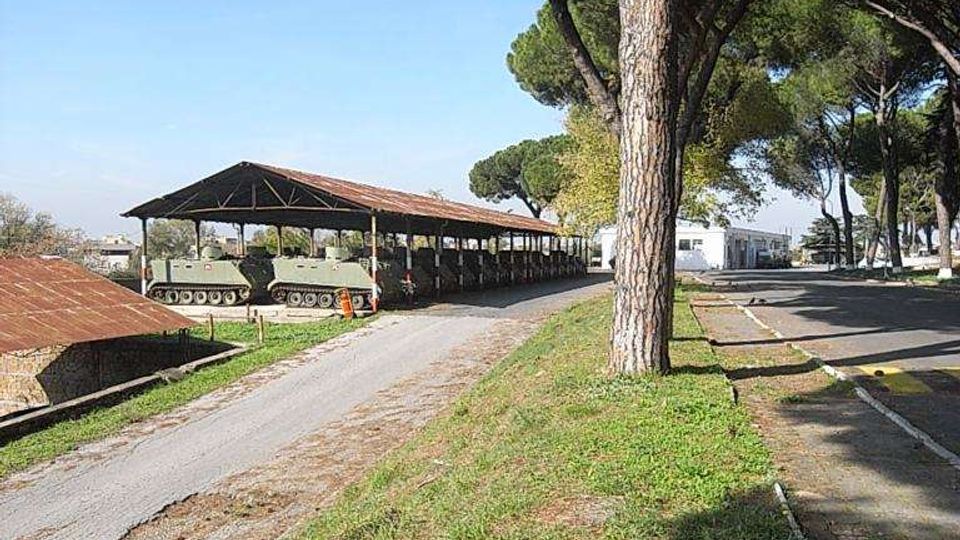 Area esterna con strada asfaltata affiancata da prato e alti pini; a sinistra si vede una lunga tettoia metallica aperta che copre veicoli cingolati. Sullo sfondo sono presenti edifici bassi e altri alberi sotto un cielo sereno.