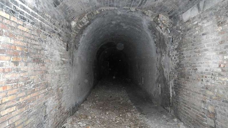 Interno di un tunnel sotterraneo in mattoni con volta a botte, pareti grezze e pavimento coperto di ghiaia e detriti. Il corridoio si restringe in prospettiva fino a un’area buia in fondo.