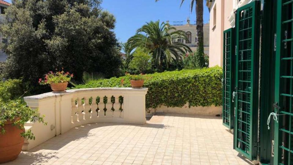 Terrazza pavimentata con ringhiera in muratura e vasi di fiori, affacciata su un giardino con siepi e palme sotto cielo sereno. A destra si vede la facciata dell’edificio con grandi porte-finestre dotate di inferriate verdi.