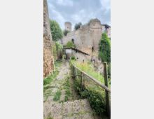 Sentiero in pietra in discesa tra alte mura di un complesso fortificato in rovina, con una torre cilindrica sullo sfondo. Ai lati si vedono una staccionata in legno e vegetazione spontanea, con un piccolo edificio addossato alle mura sotto un cielo nuvoloso.