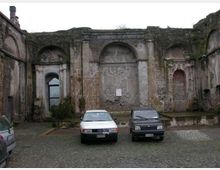 Cortile lastricato in sampietrini con una facciata in muratura antica e deteriorata, scandita da grandi nicchie ad arco e pilastri in pietra. Sullo sfondo si notano un portale ad arco con infisso vetrato e parti di pareti scrostate con tracce decorative.