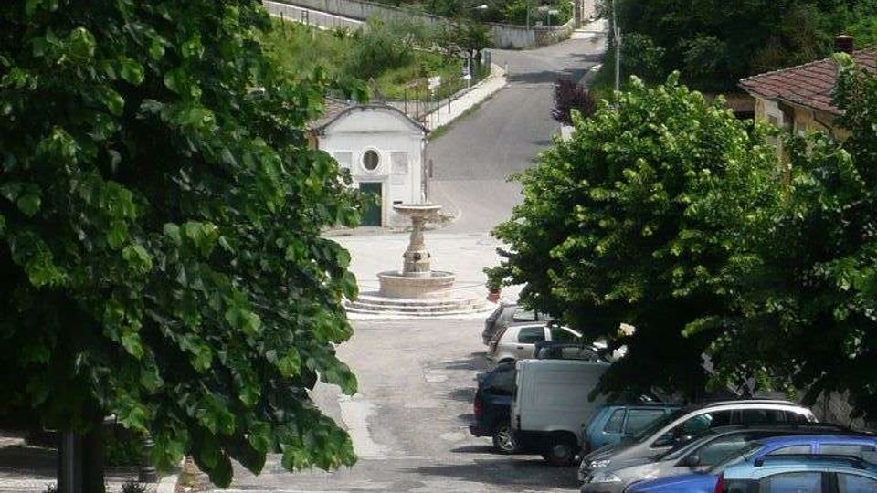 Piazza di un piccolo paese con pavimentazione in pietra, parcheggi laterali e una fontana al centro vicino a un edificio bianco. Sullo sfondo una strada sale verso case sparse su una collina verde con alberi e uliveti.