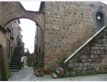 Vicolo acciottolato in un borgo storico, stretto tra edifici in pietra con archi di collegamento sopraelevati. Scale in pietra su entrambi i lati e vasi con piante lungo le pareti, con un altro arco visibile sullo sfondo.