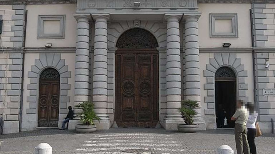Facciata di un edificio istituzionale in stile neoclassico con grande portale centrale in legno, colonne bugnate e archi laterali. Davanti c’è una piazza lastricata in sampietrini con dissuasori e vasi, e sul tetto sventolano le bandiere italiana e dell’Unione Europea.