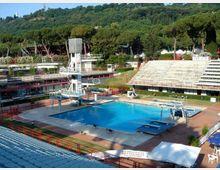 Piscina all’aperto in un complesso sportivo con vasca rettangolare azzurra, piattaforme e torre per tuffi, circondata da bordo pavimentato e ringhiere. Intorno si vedono gradinate tipo stadio e una collina boscosa sullo sfondo.