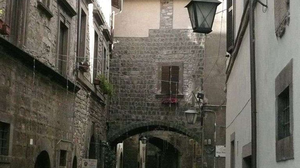 Vicolo stretto in un borgo storico, fiancheggiato da edifici in pietra e intonaco con finestre e lanterne a muro. In fondo si vede un arco in muratura che collega due edifici e, sopra, una torre in pietra.