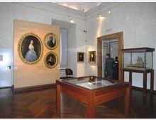 Sala museale con pareti chiare e pavimento in parquet, con grandi ritratti incorniciati appesi alle pareti e teche espositive in legno e vetro al centro e lungo il lato destro. Un’ampia apertura con cornice in legno conduce a un’altra stanza, illuminata da faretti a soffitto.