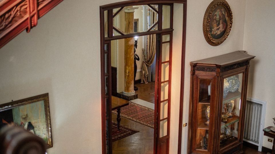 Interno di una casa storica con scala in legno e ringhiera in ferro battuto decorato, affacciata su un pianerottolo con parquet e tappeti. Sulla parete ci sono una vetrina in legno con oggetti esposti, un quadro ovale e una porta a vetri che conduce a un corridoio con colonne e tende.