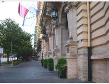 Facciata di un edificio storico in pietra con colonne e mensole scolpite, lanterne appese e bandiere esposte sopra un ampio marciapiede lastricato. Lungo la strada alberata si vedono grandi fioriere con cespugli e la carreggiata sul lato sinistro.