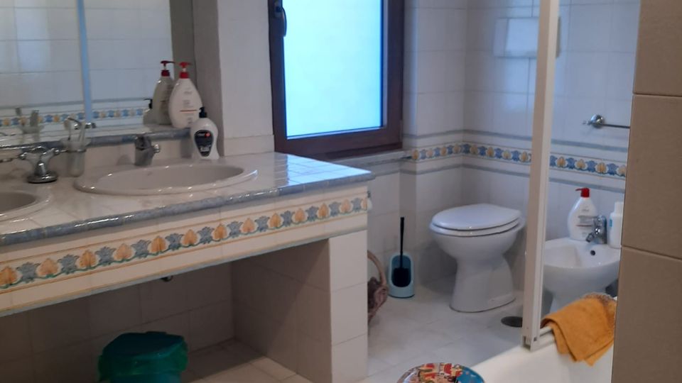 Bagno piastrellato in bianco con fascia decorativa, doppio lavabo con grande specchio e finestra con telaio in legno. A destra si vedono WC e bidet e una vasca con parete in vetro, con alcuni oggetti appoggiati sul bordo.