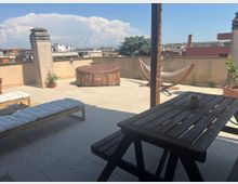 Terrazza sul tetto pavimentata con parapetto basso, arredata con tavolo e panche in legno, lettini, una vasca idromassaggio rotonda e un’amaca su supporto in legno. Sullo sfondo si vede il panorama dei tetti e degli edifici circostanti sotto un cielo sereno con grandi nuvole.
