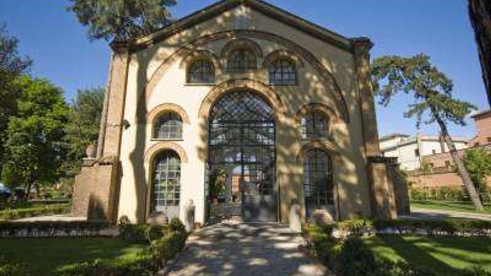 Facciata di un edificio storico con grande ingresso ad arco vetrato centrale e finestre ad arco, raggiunto da un vialetto in pietra. L’edificio è circondato da prato, aiuole e alti alberi in un’area verde.