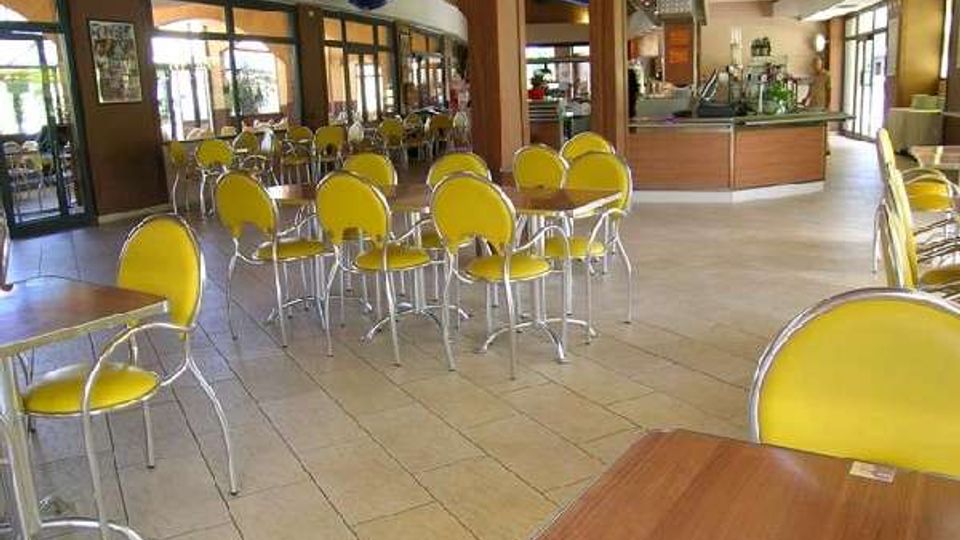 Interno di un bar o caffetteria con pavimento in piastrelle, tavolini in legno e sedie gialle disposte nell’area centrale. Sullo sfondo si vede un bancone curvo in legno con attrezzature, colonne portanti e grandi vetrate che danno su una zona esterna coperta.