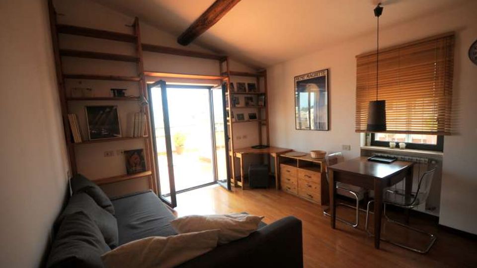 Interno di un soggiorno con pavimento in legno, divano grigio e scaffalature a parete con scala e travi a vista sul soffitto. Una porta finestra apre su un balcone luminoso, mentre a destra ci sono un tavolo con sedie, una lampada a sospensione e una finestra con veneziane.