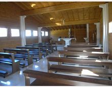 Interno di una piccola cappella in legno con soffitto a travi, file di panche scure e finestre rettangolari laterali che lasciano entrare luce naturale. In fondo si vede l’altare bianco con crocifisso, sostenuto da colonne bianche lungo la navata.