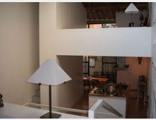 Interno di una casa moderna su più livelli con soppalco e parapetti bianchi, soffitto con travi in legno a vista e pavimento in legno. In basso si vede una cucina open space con isola e zona pranzo sullo sfondo, illuminata da lampade con paralume bianco.