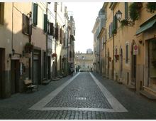Strada stretta in pavé con marciapiedi in pietra, fiancheggiata da edifici storici a più piani con persiane, balconcini e lanterne a muro. Lungo le facciate si vedono ingressi di negozi, panchine e piante in vaso, con la strada che termina verso un edificio sullo sfondo.