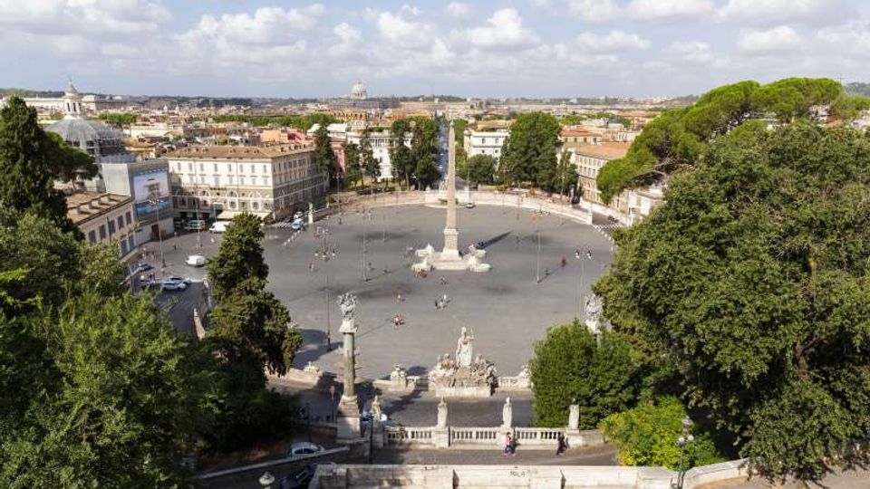 Veduta dall’alto di una grande piazza circolare con un alto obelisco al centro e fontane monumentali alla base, circondata da edifici storici. Ai margini si notano ampie aree alberate e, sullo sfondo, il profilo urbano con cupole e tetti sotto un cielo parzialmente nuvoloso.