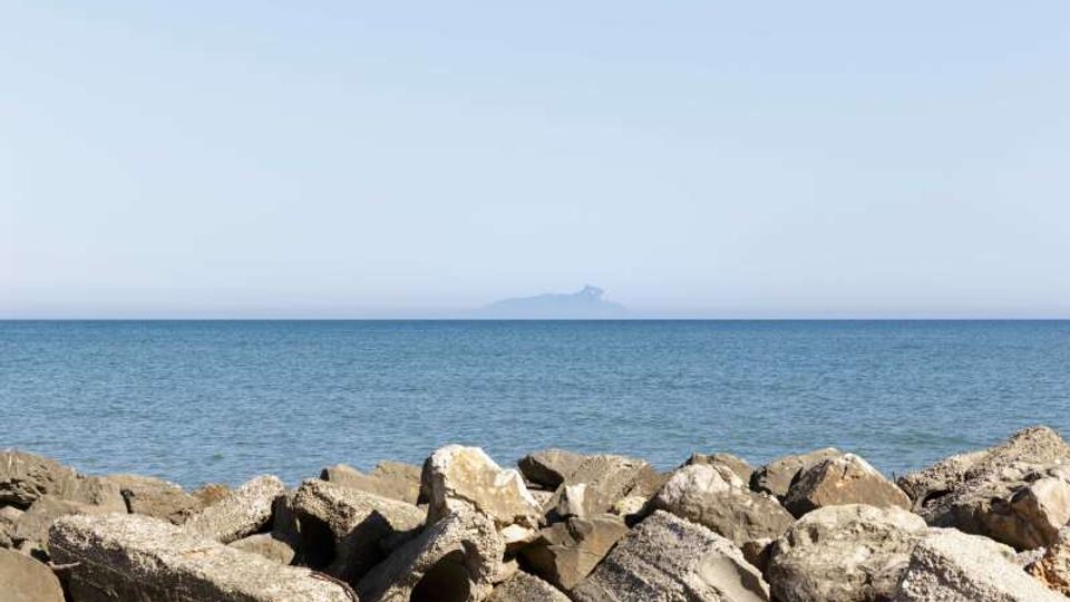 Costa marina con una barriera di grandi massi e blocchi di cemento in primo piano, affacciata su un mare calmo con orizzonte netto. In lontananza si intravede una piccola isola o promontorio sullo sfondo sotto un cielo sereno.