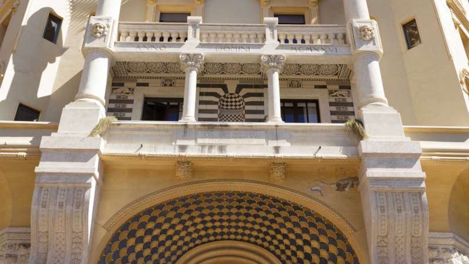 Facciata di un edificio storico a più piani vista dal basso, con ampio portale ad arco decorato a mosaico, scalinata d’ingresso e balconi sovrapposti. La struttura presenta colonne, logge ad arcate e tetto sporgente con elementi ornamentali in pietra.