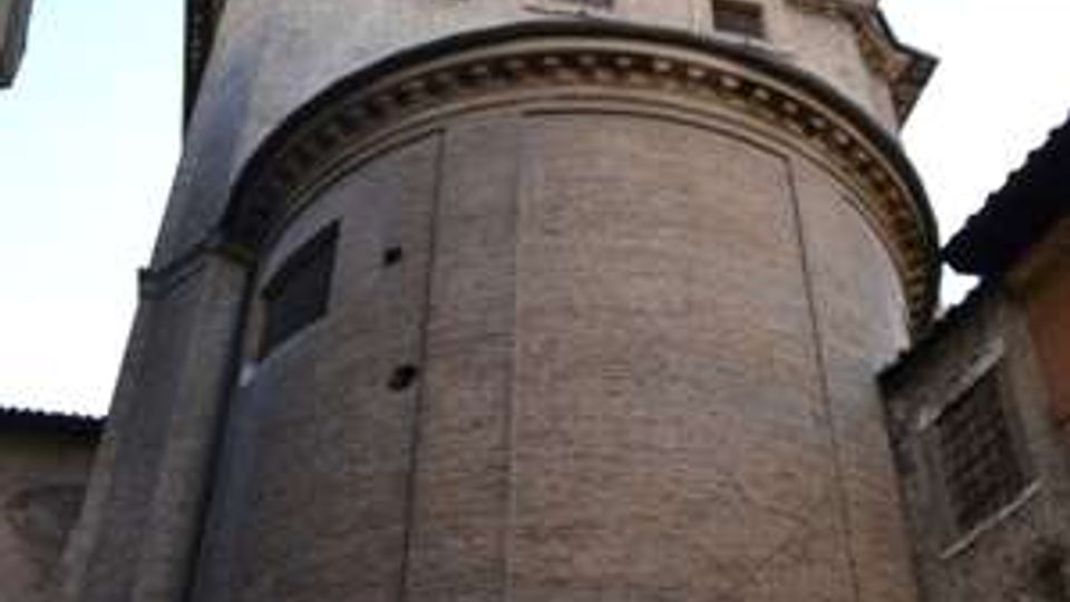 Abside semicircolare in mattoni di una chiesa storica, con cornice decorativa sotto la gronda e piccole finestre nella parte superiore. Inquadratura dal basso in una stretta via tra edifici, con cielo sereno sullo sfondo.