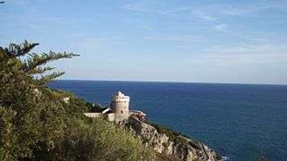 Torre costiera in pietra su un promontorio roccioso, circondata da vegetazione mediterranea, con il mare aperto sullo sfondo sotto un cielo sereno.