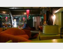 Interno di un locale tipo lounge con soffitto industriale e illuminazione soffusa, arredato con grandi divani e pouf arancioni. Nello spazio si vedono tendaggi colorati e pannelli divisori, con un bancone chiaro sulla destra e luci decorative sullo sfondo.